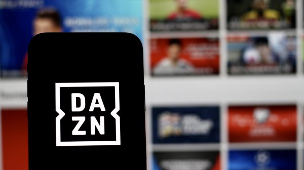 FootballR - NFL - Ein Smartphone mit dem DAZN-Logo auf dem Bildschirm wird vor einen unscharfen Hintergrund gehalten. Der Hintergrund scheint ein Computer- oder Fernsehbildschirm zu sein, auf dem verschiedene sportbezogene Miniaturansichten angezeigt werden, darunter NFL Game Pass Inhalte, wodurch der digitale Streaming-Kontext im Zusammenhang mit DAZN hervorgehoben wird. Diese Beschreibung wurde automatisch generiert.