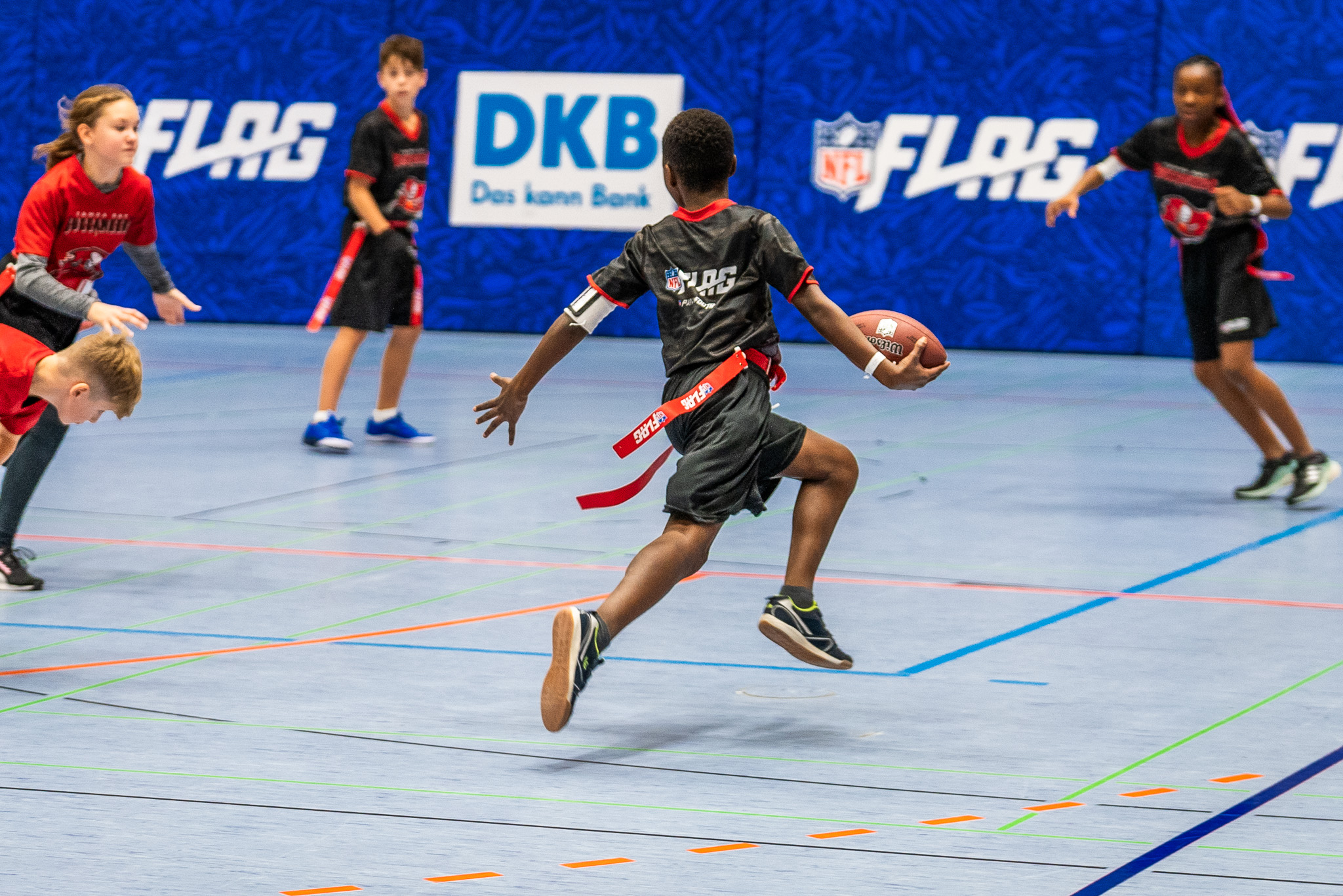FootballR - NFL - Ein kleiner Junge in schwarzer Sportkleidung springt während eines Flag-Football-Spiels in der Saison 2024 mit einem Football in die Luft. Er ist von anderen Kindern umgeben, manche in roten und manche in schwarzen Uniformen, auf einem blauen Hallenplatz, im Hintergrund mit dem NFL-FLAG-Logo und DKB-Schildern geschmückt. Diese Beschreibung wurde automatisch generiert.