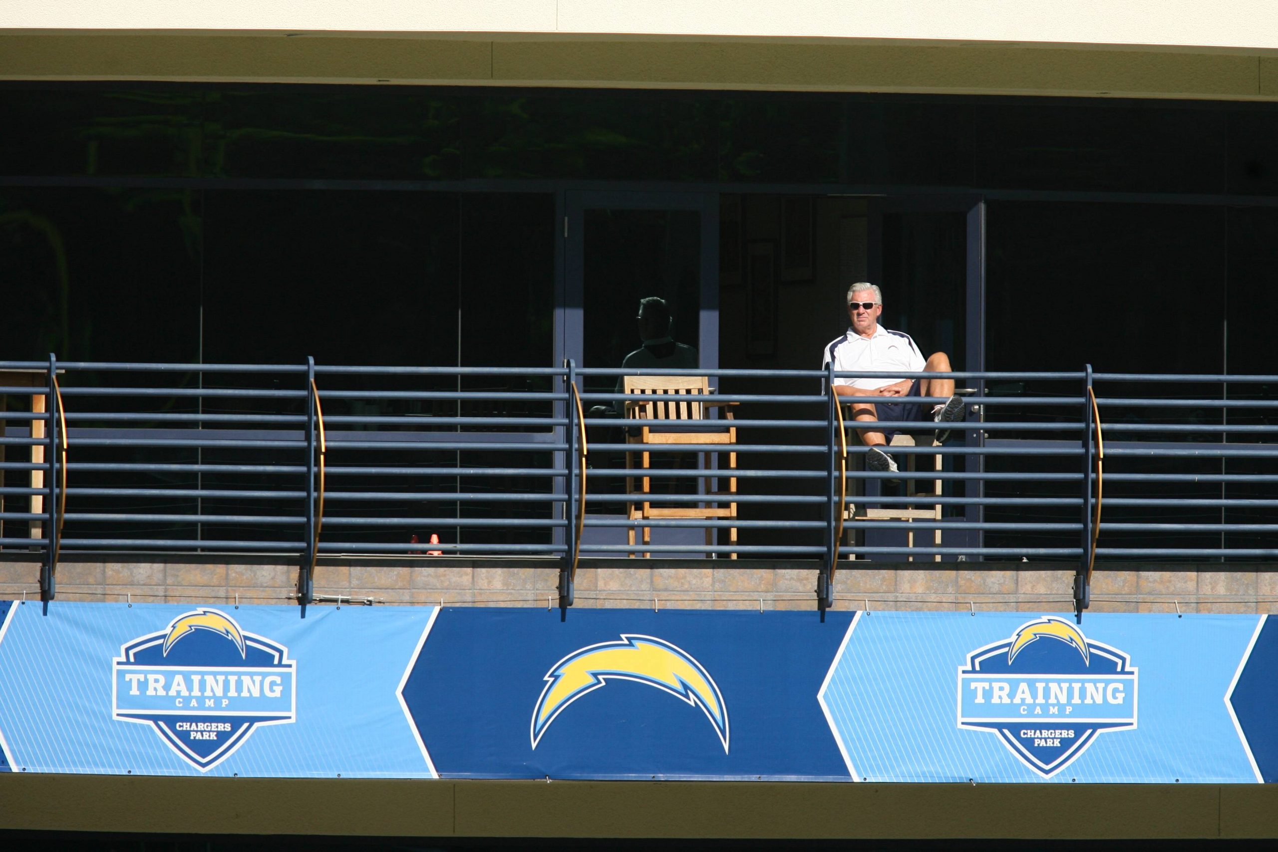 FootballR - NFL A.J. Smith - Ein Mann mit Sonnenbrille und weißem Poloshirt sitzt hinter einem Geländer mit dem Logo des NFL-Teams der Chargers. Er wirkt entspannt und beobachtet das Geschehen an einem sonnigen Tag von einem schattigen Balkon aus. Im Hintergrund mischen sich die General Manager verschiedener NFL-Teams untereinander. Diese Beschreibung wurde automatisch generiert.