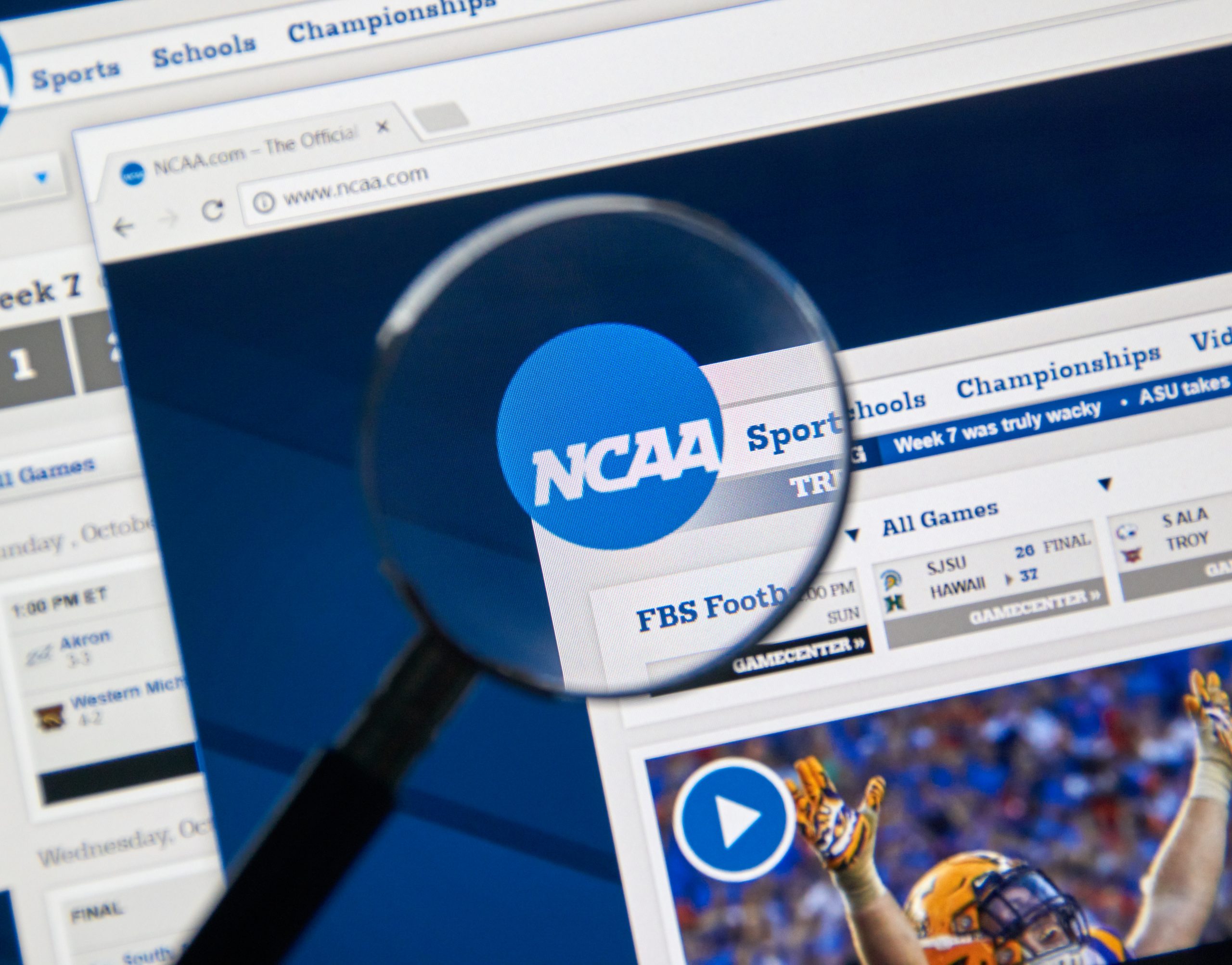 FootballR - NFL - Eine Lupe konzentriert sich auf das NCAA Logo, das auf einem Computerbildschirm angezeigt wird, und hebt in den Browser-Tabs verschiedene Abschnitte wie „Sport“, „Schulen“ und „Meisterschaften“ hervor. Unter dem Logo gibt es einen Zeitplan für Footballspiele und ein Video-Thumbnail mit einem Spieler in orange-weißer Uniform, was die umfangreichen Kommerzialisierungsbemühungen der NCAA hervorhebt. Diese Beschreibung wurde automatisch generiert.