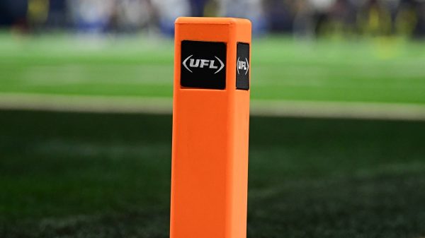 FootballR - NFL - Nahaufnahme eines orangefarbenen Pylons mit dem UFL -Logo auf einem Footballfeld. Der grüne Rasen ist sichtbar, mit weißen und schwarzen Feldmarkierungen. Im Hintergrund sind verschwommene Spieler in Uniformen zu sehen. Der Fokus liegt auf dem Pylon, der die Textur und die Logos hervorhebt und eine weitere erfolgreiche Saison für Zuseher symbolisiert. Diese Beschreibung wurde automatisch generiert.
