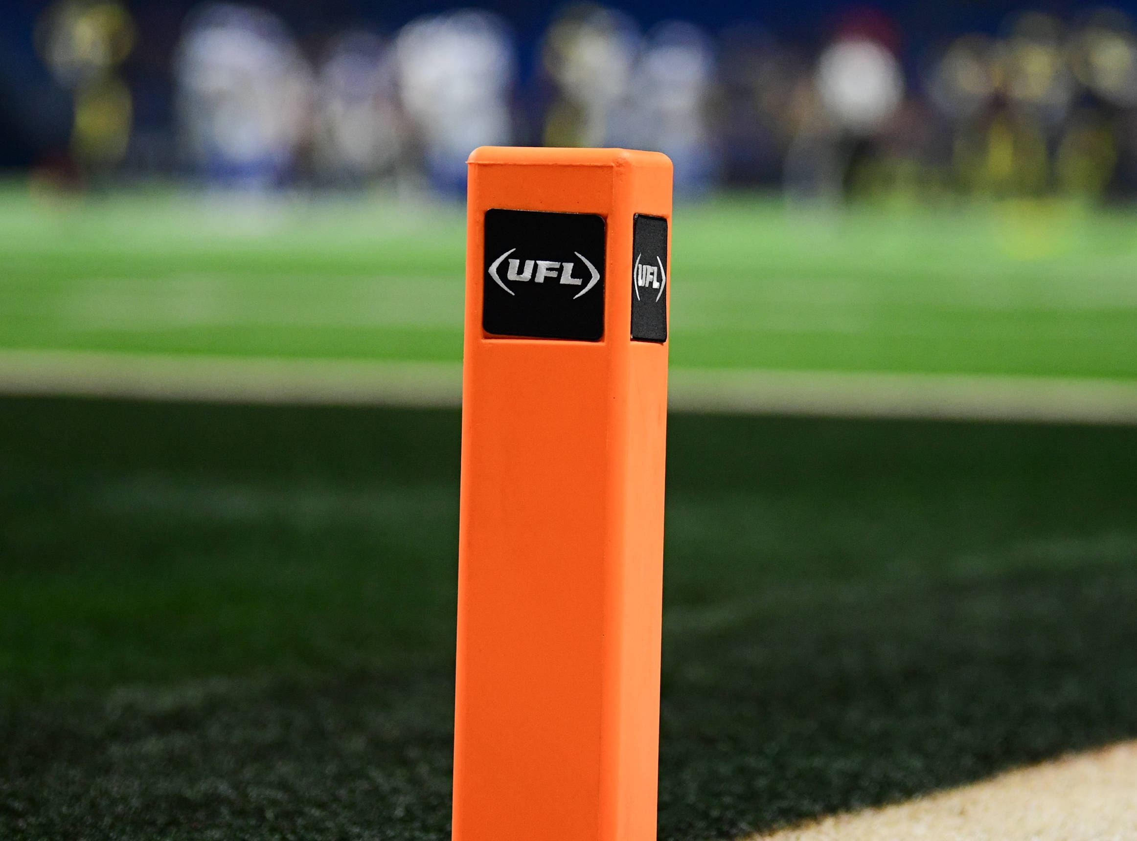 FootballR - NFL - Nahaufnahme eines orangefarbenen Pylons mit dem UFL -Logo auf einem Footballfeld. Der grüne Rasen ist sichtbar, mit weißen und schwarzen Feldmarkierungen. Im Hintergrund sind verschwommene Spieler in Uniformen zu sehen. Der Fokus liegt auf dem Pylon, der die Textur und die Logos hervorhebt und eine weitere erfolgreiche Saison für Zuseher symbolisiert. Diese Beschreibung wurde automatisch generiert.