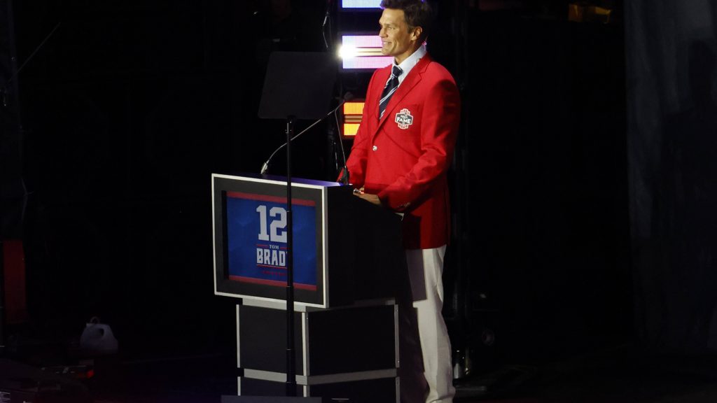 FootballR - NFL - Ein Mann steht lächelnd an einem Podium und trägt einen leuchtend roten Blazer und weiße Hosen. Auf dem Podium sind die Nummer 12 und das Wort „BRADY“ zu sehen. Im schwach beleuchteten Hintergrund mit bunten vertikalen Lichtern ist klar, dass er die Patriots-Legende Tom Brady geehrt wird. Diese Beschreibung wurde automatisch generiert.