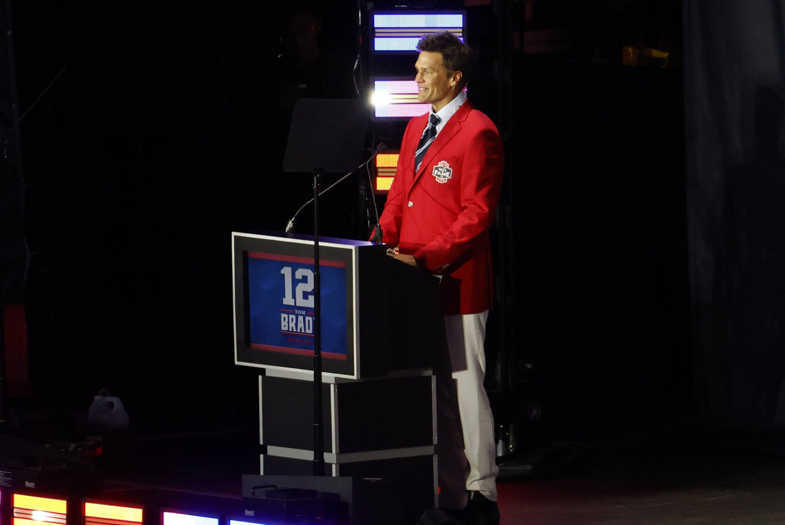 FootballR - NFL - Ein Mann steht lächelnd an einem Podium und trägt einen leuchtend roten Blazer und weiße Hosen. Auf dem Podium sind die Nummer 12 und das Wort „BRADY“ zu sehen. Im schwach beleuchteten Hintergrund mit bunten vertikalen Lichtern ist klar, dass er die Patriots-Legende Tom Brady geehrt wird. Diese Beschreibung wurde automatisch generiert.