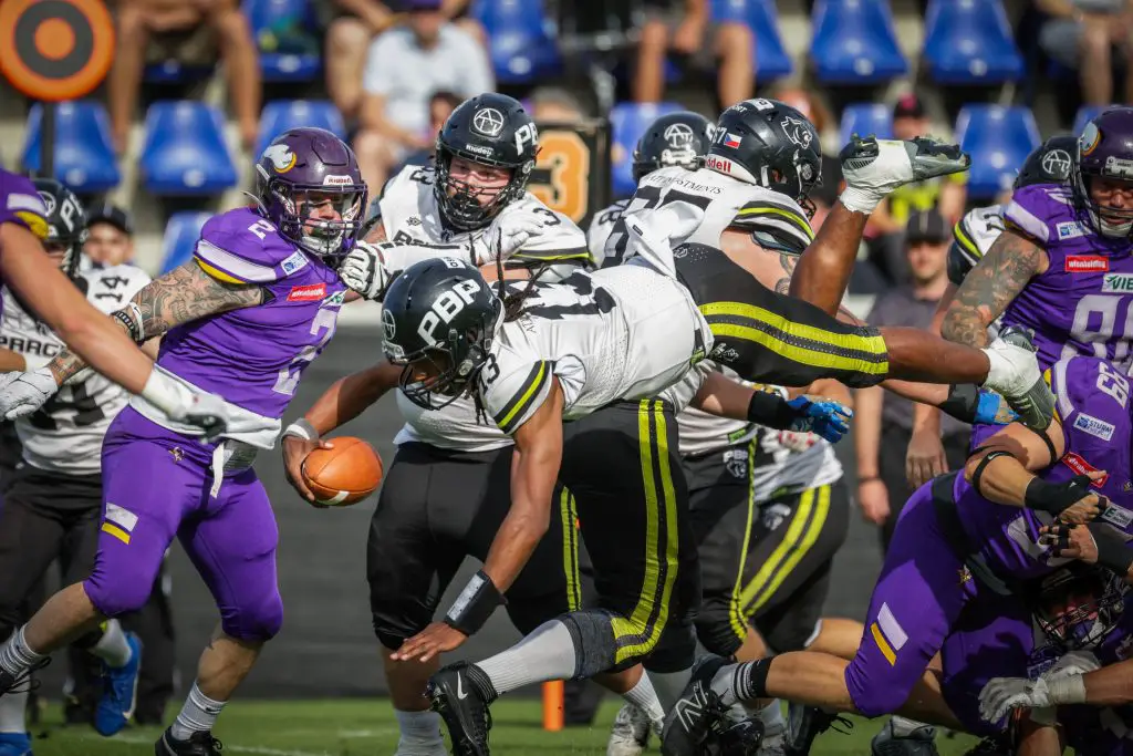 FootballR - NFL - Ein Footballspieler in weißem Trikot und schwarzer Hose hechtet mit dem Ball in der Hand über das Spielfeld und zielt auf einen Touchdown beim Austrian Bowl XXXIX. Er ist in der Luft und Verteidiger in lila Trikots versuchen, ihn zu tackeln. Andere Spieler in weißen und schwarzen Trikots blockieren die Verteidiger. Im Hintergrund sind Zuschauer auf blauen Sitzen zu sehen. Diese Beschreibung wurde automatisch generiert.