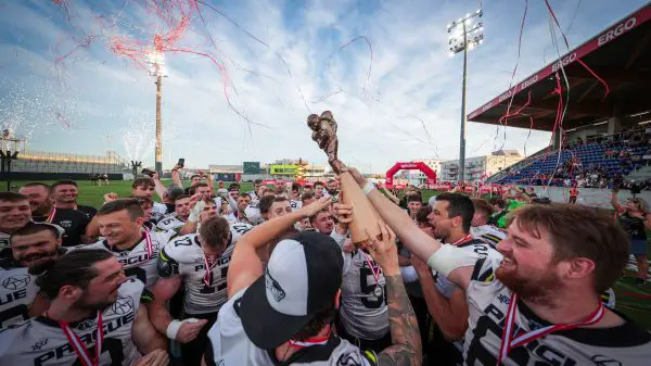 FootballR - NFL - Eine jubelnde Football-Mannschaft in weiß-schwarzen Trikots mit der Aufschrift „PREGLUE“ auf der Brust reckt unter einem klaren blauen Himmel gemeinsam den Pokal des Austrian Bowl XXXIX-Siegers in die Höhe. Rotes Konfetti und Luftschlangen liegen in der Luft, während sie auf einem gut beleuchteten Footballfeld feiern, im Hintergrund sind Zuschauerränge zu sehen. Diese Beschreibung wurde automatisch generiert.