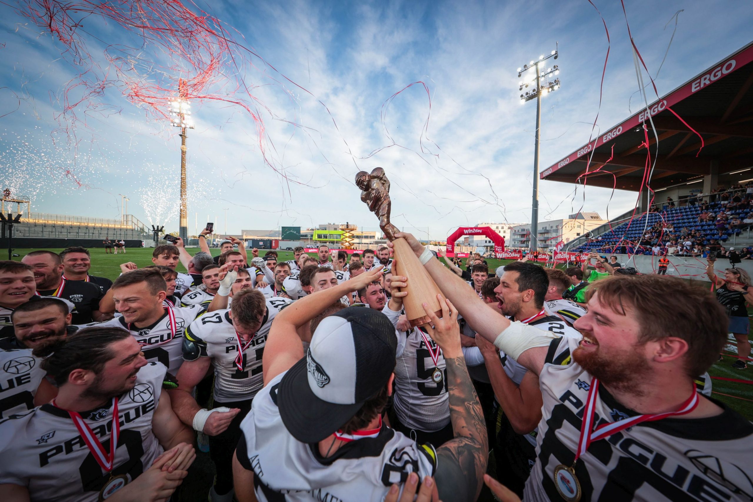 FootballR - NFL - Eine jubelnde Football-Mannschaft in weiß-schwarzen Trikots mit der Aufschrift „PREGLUE“ auf der Brust reckt unter einem klaren blauen Himmel gemeinsam den Pokal des Austrian Bowl XXXIX-Siegers in die Höhe. Rotes Konfetti und Luftschlangen liegen in der Luft, während sie auf einem gut beleuchteten Footballfeld feiern, im Hintergrund sind Zuschauerränge zu sehen. Diese Beschreibung wurde automatisch generiert.