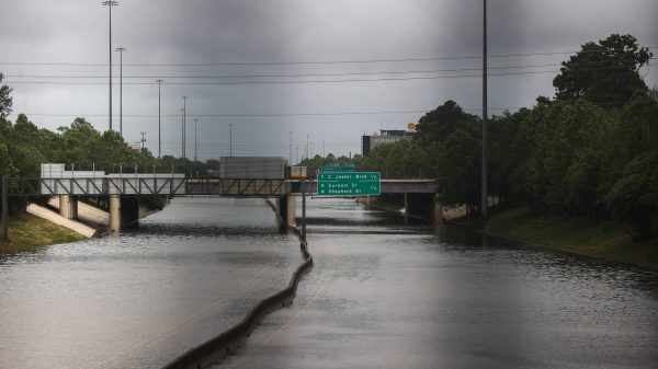 FootballR - NFL - Eine Autobahn in Houston ist stark überflutet. Das Wasser reicht bis zu den Verkehrsschildern und überschwemmt sie teilweise. Die Autobahnschilder über der Straße zeigen Richtungen und Entfernungen zur Innenstadt und zu wichtigen Ausfahrten wie dem NRG Stadium an. Der Himmel ist bedeckt, was auf einen kürzlichen Sturm hindeutet, möglicherweise durch Hurrikan Beryl, während Bäume die Seiten der Autobahn säumen. Diese Beschreibung wurde automatisch generiert.