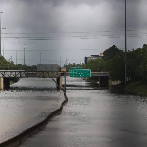 FootballR - NFL - Eine Autobahn in Houston ist stark überflutet. Das Wasser reicht bis zu den Verkehrsschildern und überschwemmt sie teilweise. Die Autobahnschilder über der Straße zeigen Richtungen und Entfernungen zur Innenstadt und zu wichtigen Ausfahrten wie dem NRG Stadium an. Der Himmel ist bedeckt, was auf einen kürzlichen Sturm hindeutet, möglicherweise durch Hurrikan Beryl, während Bäume die Seiten der Autobahn säumen. Diese Beschreibung wurde automatisch generiert.