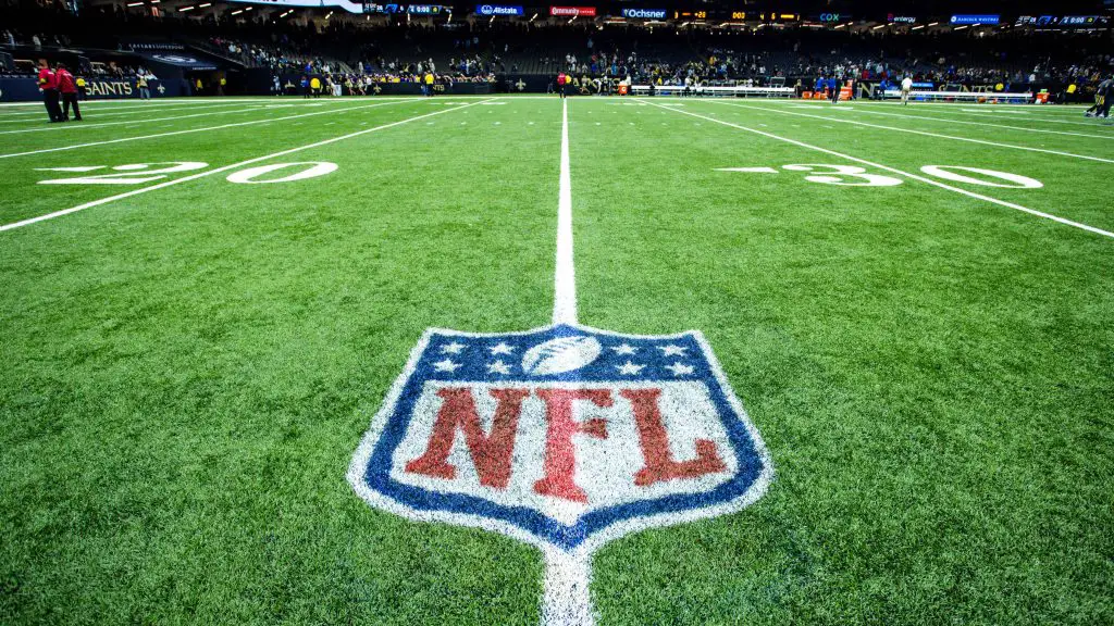 FootballR - NFL - Ein Footballfeld mit dem NFL-Logo in der Mitte zwischen der 20- und 30-Yard-Linie. Das Stadion ist beleuchtet und im Hintergrund voller Zuschauer. Eine Anzeigetafel zeigt „FINAL STATS“ für die Saison 2023 an. Das Feld hat deutlich erkennbare weiße Yard-Linien und Hash-Markierungen, was auf eine gut gepflegte Spielfläche hinweist. Diese Beschreibung wurde automatisch generiert.