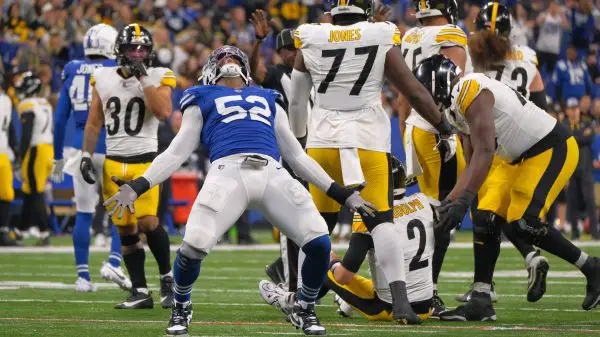 FootballR - NFL - Samson Ebukam, in seiner blauen Uniform mit der Nummer 52, steht mit ausgestreckten Armen und nach hinten geneigtem Kopf da, um zu jubeln. Spieler in weißen und gelben Uniformen reagieren auf das Spiel, einige knien oder stehen. Das Spielfeld ist mit weißen Linien markiert, während die Menge als verschwommener Hintergrund bleibt. Diese Beschreibung wurde automatisch generiert.