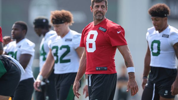 FootballR - NFL - Aaron Rodgers in einem roten Trainings-Footballtrikot mit der Nummer 8 steht auf einem Feld. Im Hintergrund sind andere Spieler zu sehen, die weiße Trikots mit grünen Nummern tragen, entweder stehend oder gehend. Der Mann ähnelt Aaron Rodgers beim Training, blickt leicht nach links und in der Ferne ist ein Gebäude zu sehen. Diese Beschreibung wurde automatisch generiert.