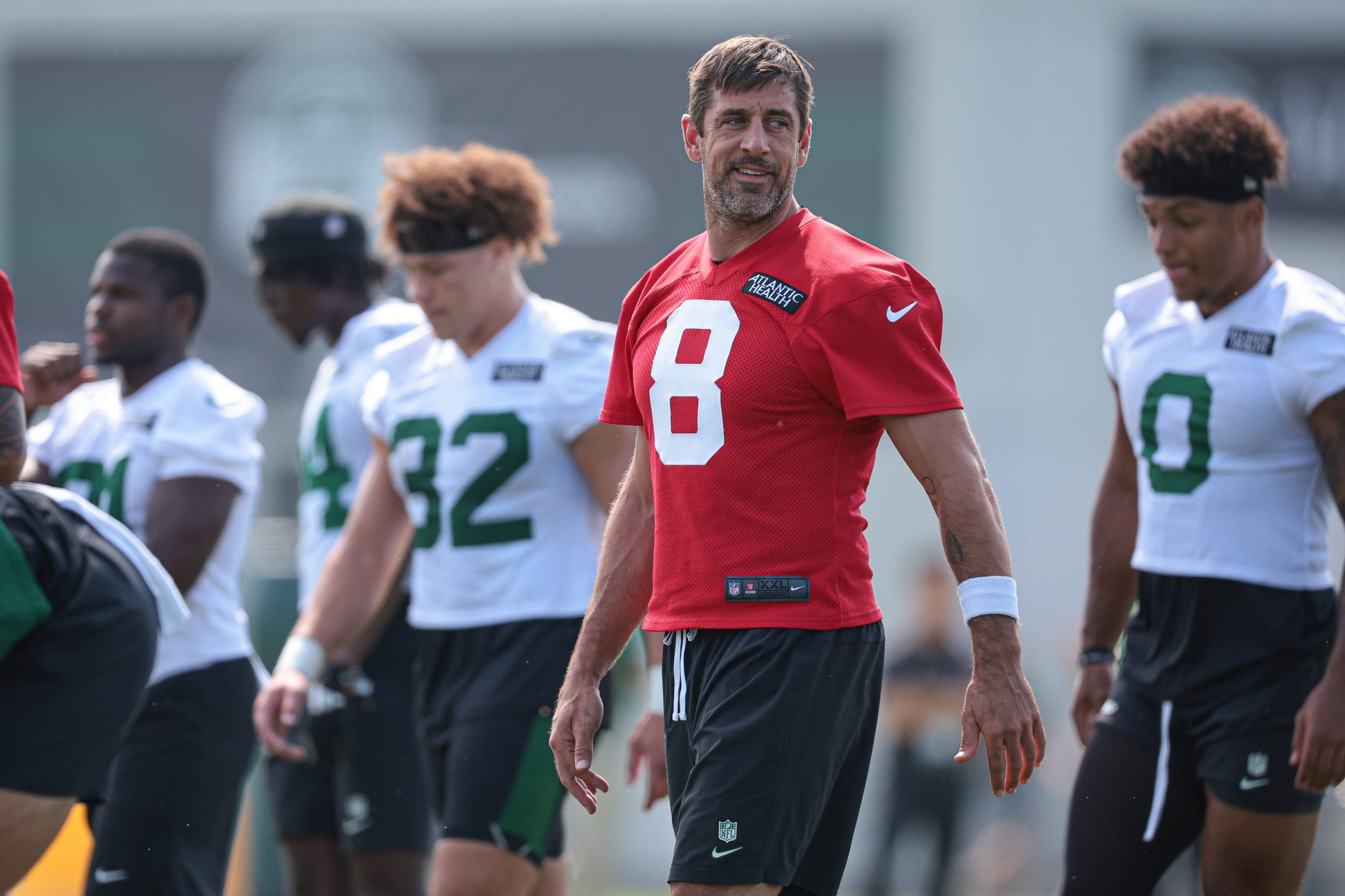 FootballR - NFL - Aaron Rodgers in einem roten Trainings-Footballtrikot mit der Nummer 8 steht auf einem Feld. Im Hintergrund sind andere Spieler zu sehen, die weiße Trikots mit grünen Nummern tragen, entweder stehend oder gehend. Der Mann ähnelt Aaron Rodgers beim Training, blickt leicht nach links und in der Ferne ist ein Gebäude zu sehen. Diese Beschreibung wurde automatisch generiert.