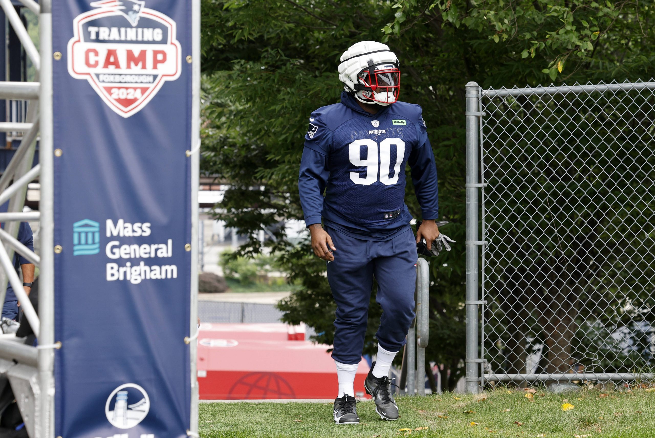 FootballR - NFL Christian Barmore - Ein Footballspieler, der ein marineblaues Patriots-Trikot mit der Nummer 90, einen weißen Helm und einen roten Gesichtsschutz trägt, läuft auf einer Rasenfläche in der Nähe eines Maschendrahtzauns. Der Spieler ist Teil eines Trainingslagers, was durch ein Banner mit den Aufschriften „Trainingslager 2024“ und „Mass General Brigham“ gekennzeichnet ist. Im Hintergrund sind Bäume zu sehen. Diese Beschreibung wurde automatisch generiert.